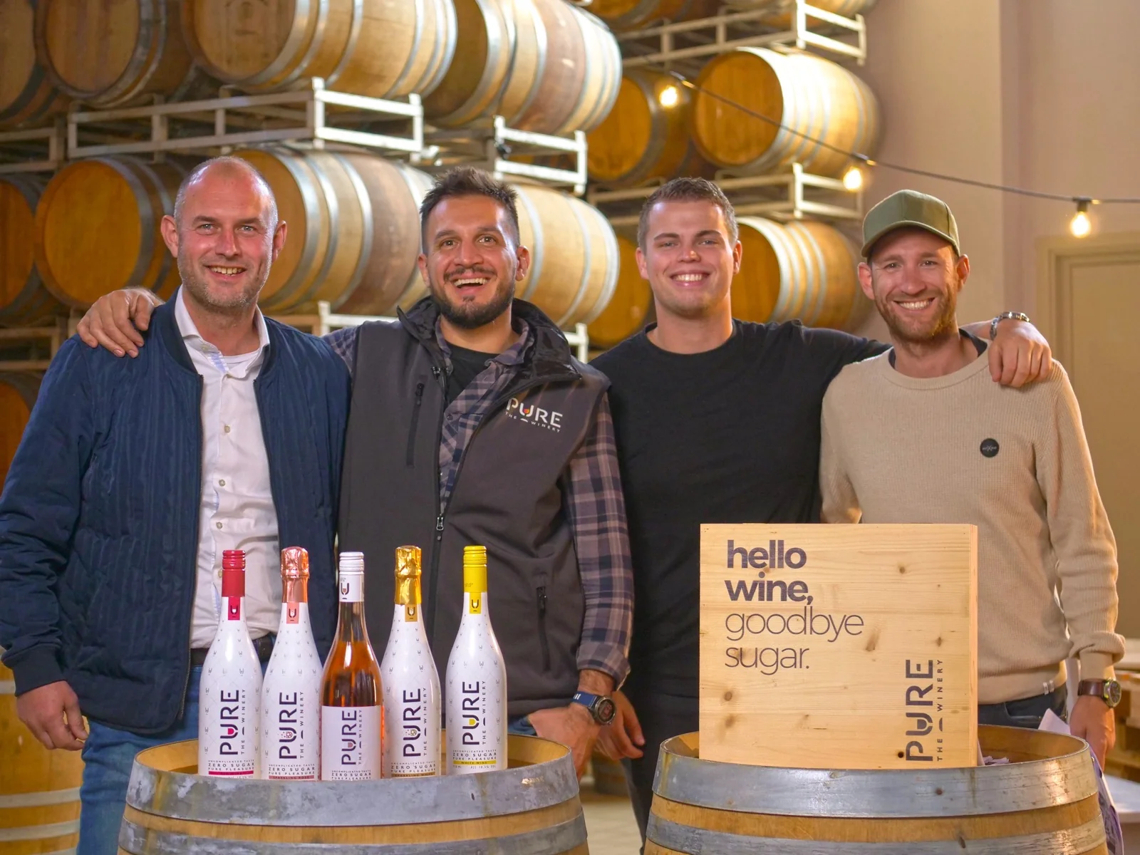 Groepsportret tussen de wijnvaten met de vijf PURE flessen en het "hello wine, goodbye sugar" houten bord
